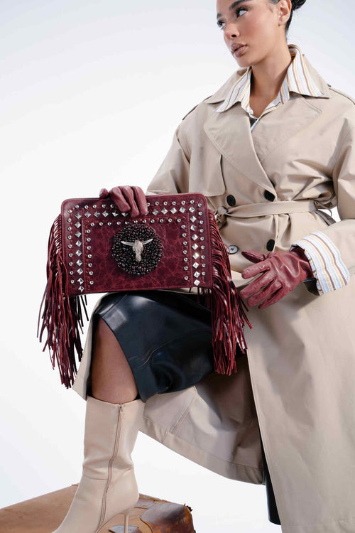 Woman holding a studded handbag with fringe details, wearing a beige trench coat and high boots.