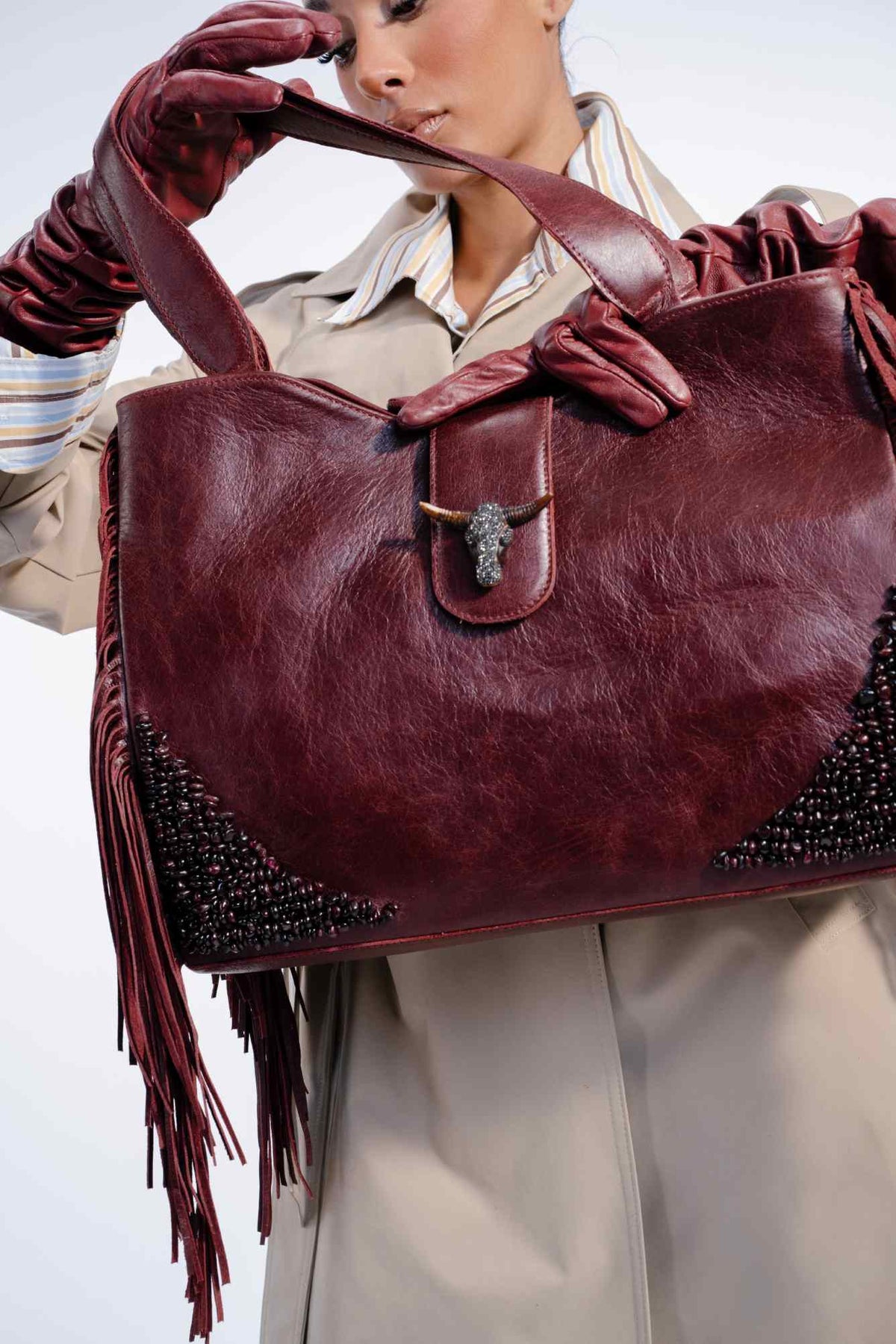 Person holding a burgundy leather handbag with fringe details on a light background