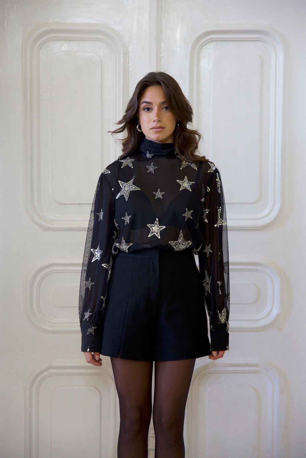 Woman wearing a black turtleneck with star patterns and black shorts against a white paneled wall cultmia.