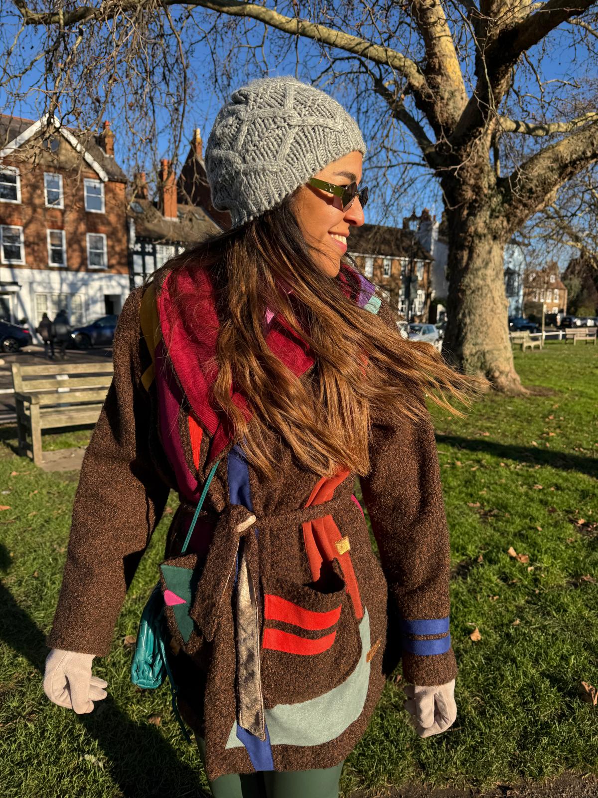 Person wearing a colorful coat and gray knit hat in an outdoor setting with trees and buildings in the background.